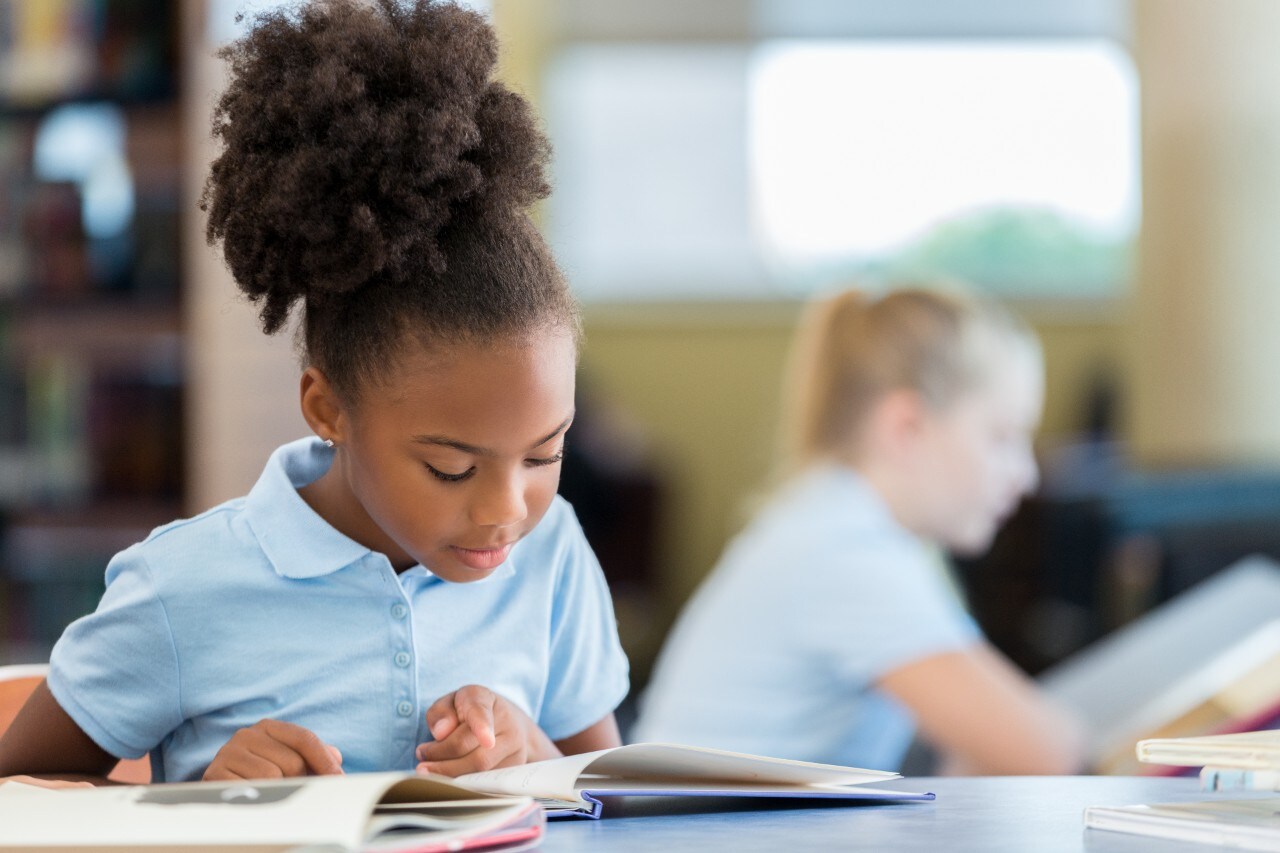 Adorable African American elementary age schoolgirl sitting at a table in the library and reading a book. Cute young African American girl reading a book at the school library.